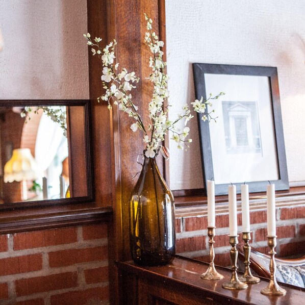 Un jarrón marrón oscuro con flores blancas sobre una repisa de madera, un cuadro enmarcado y velas blancas.