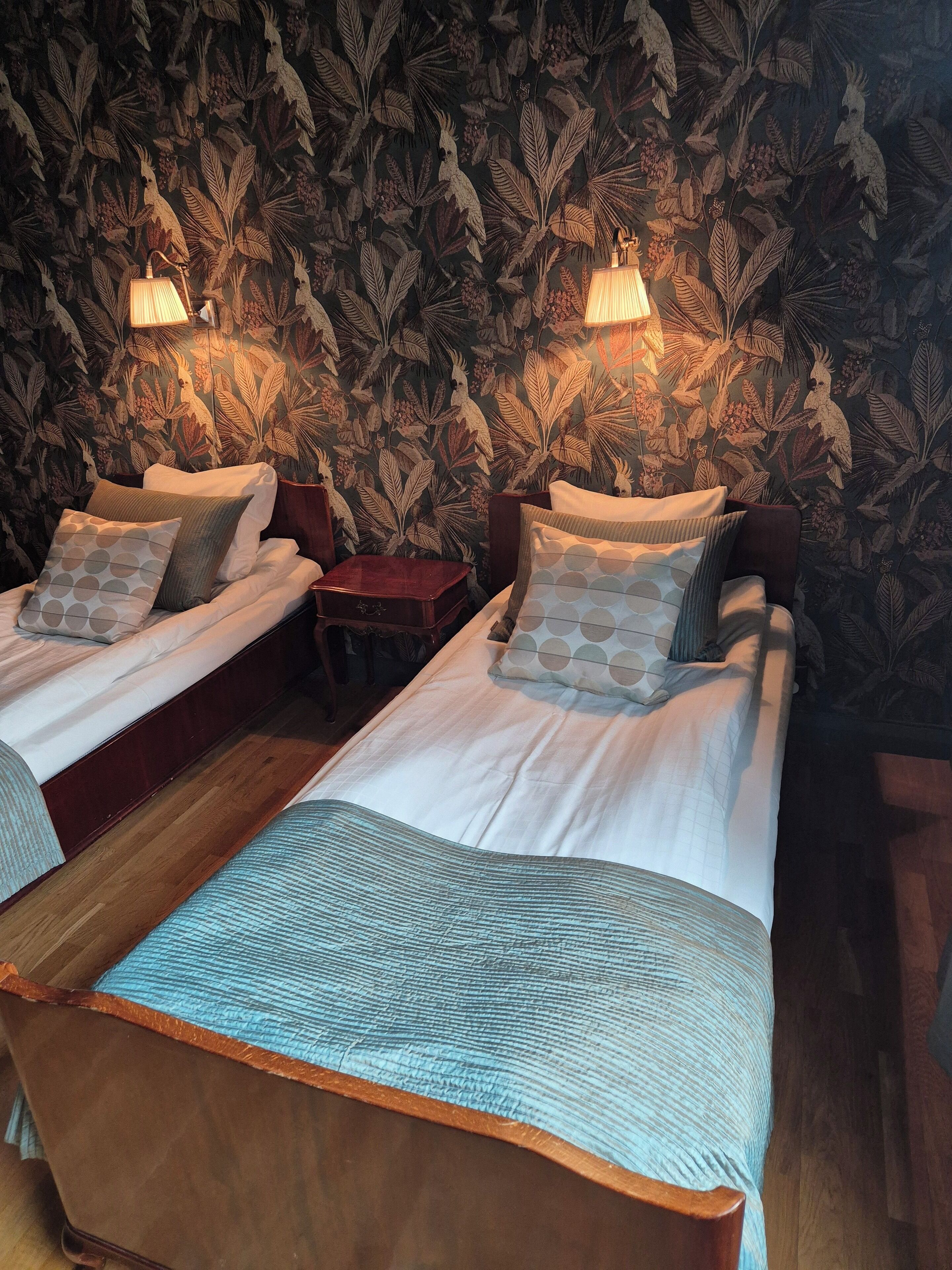 A room with two single beds, a small wooden nightstand, and a wall with a leaf pattern.