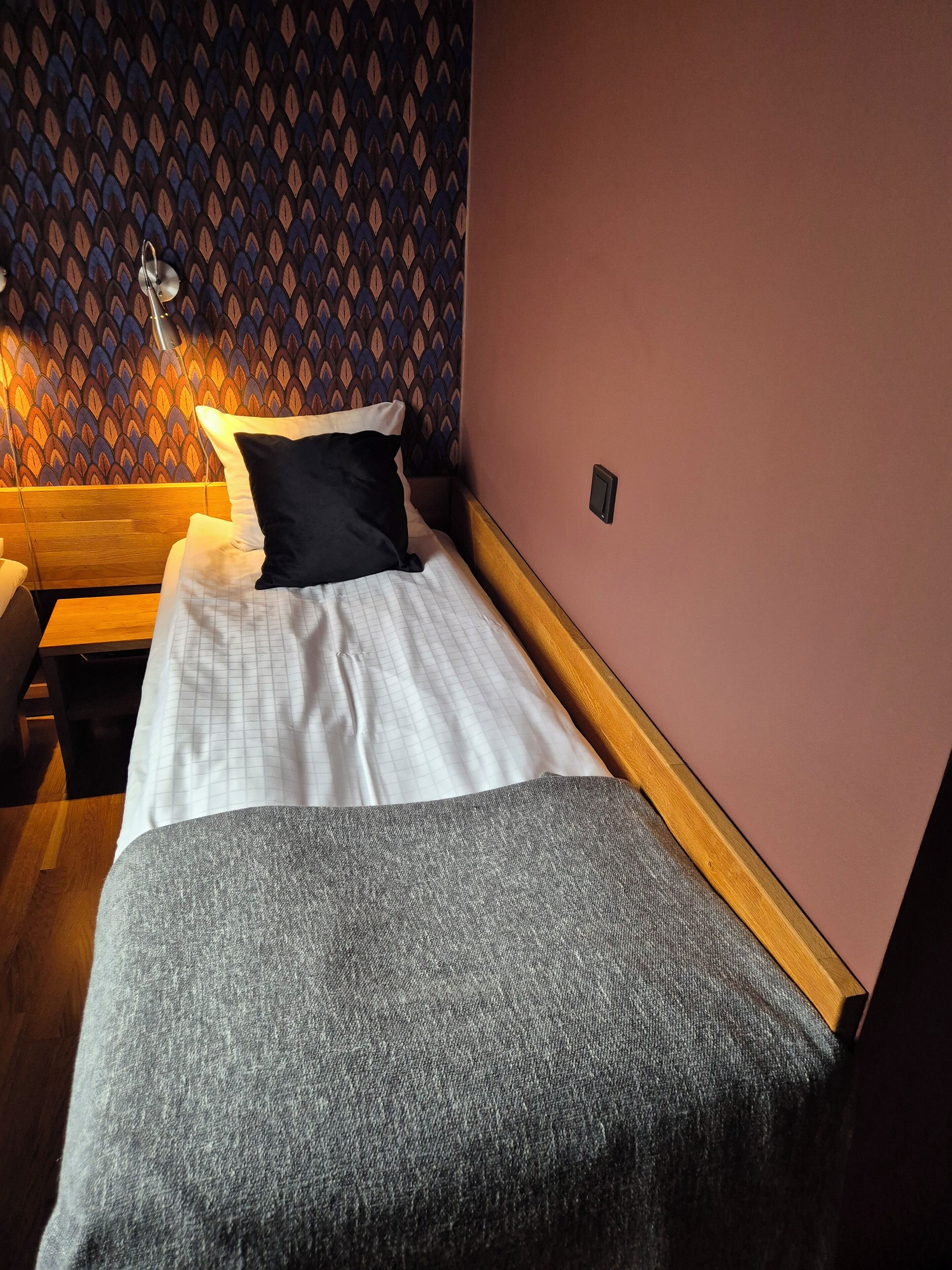 Una cama individual con una colcha blanca y gris, un almohadón negro y un cabecero de madera.