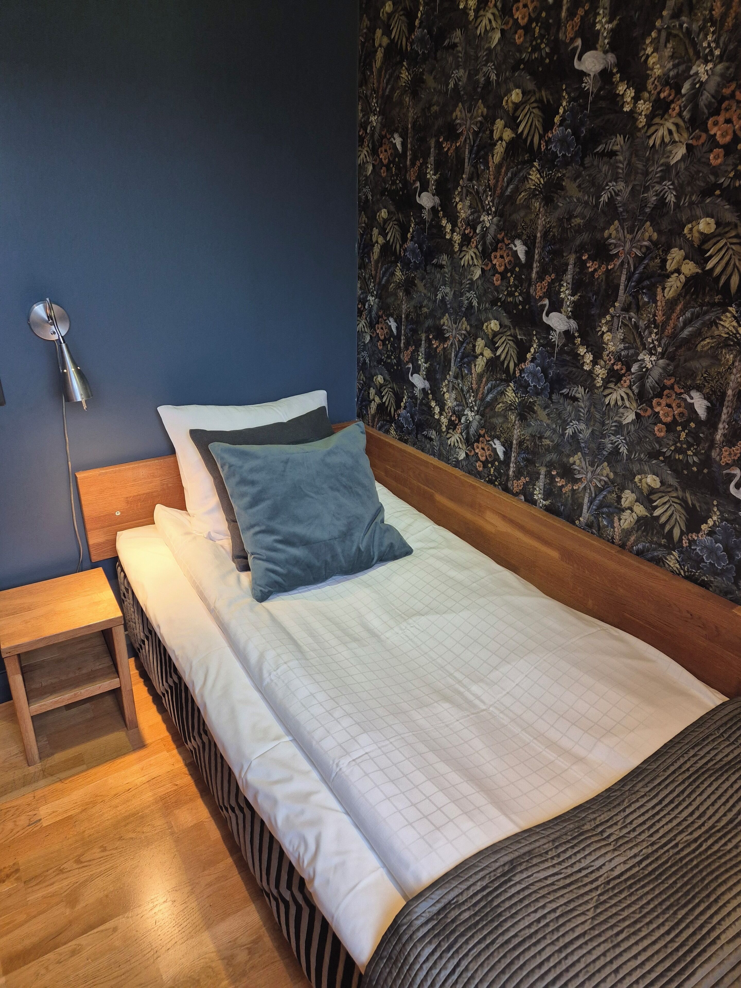 A neatly made bed with a blue pillow, a wooden bedside table, and a wall with a floral pattern.