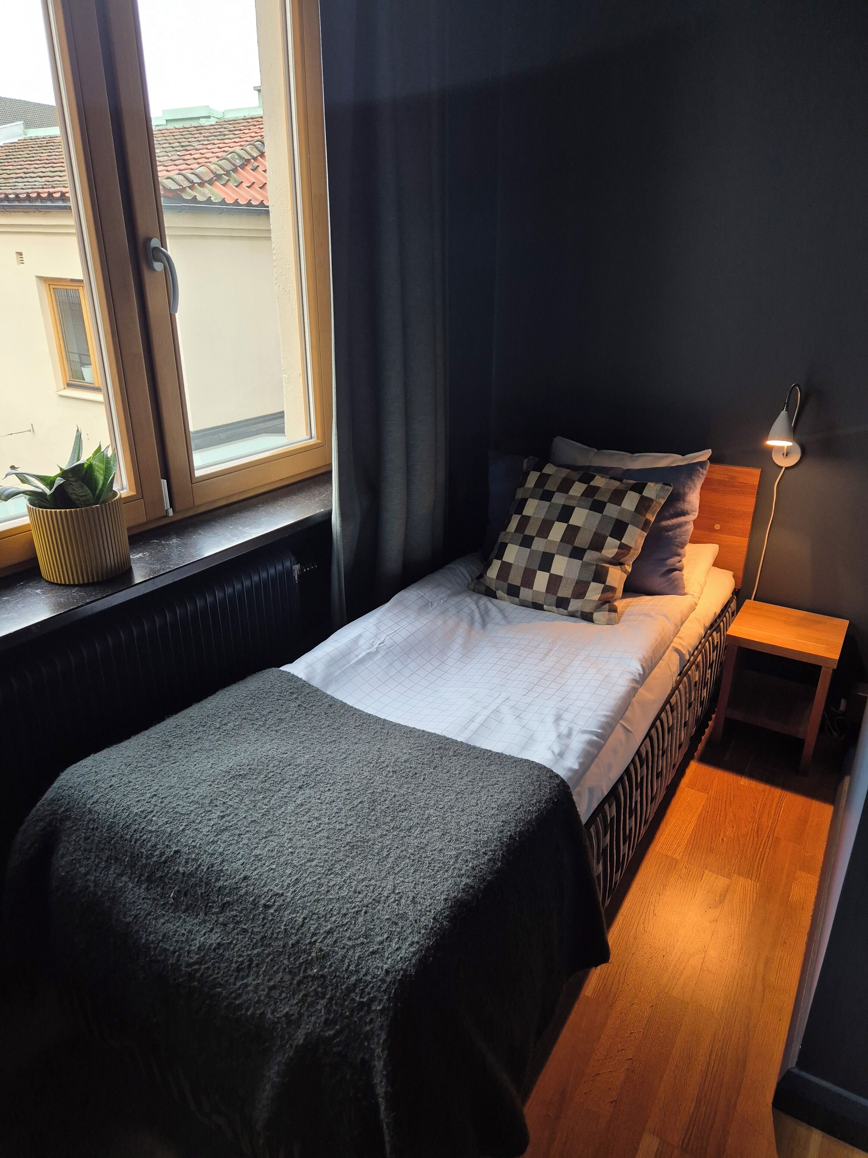 A bedroom with a bed, a bedside table with a lamp, a window with curtains, and a potted plant on the windowsill.