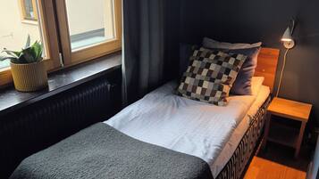 A bedroom with a bed, a bedside table with a lamp, a window with curtains, and a potted plant on the windowsill.