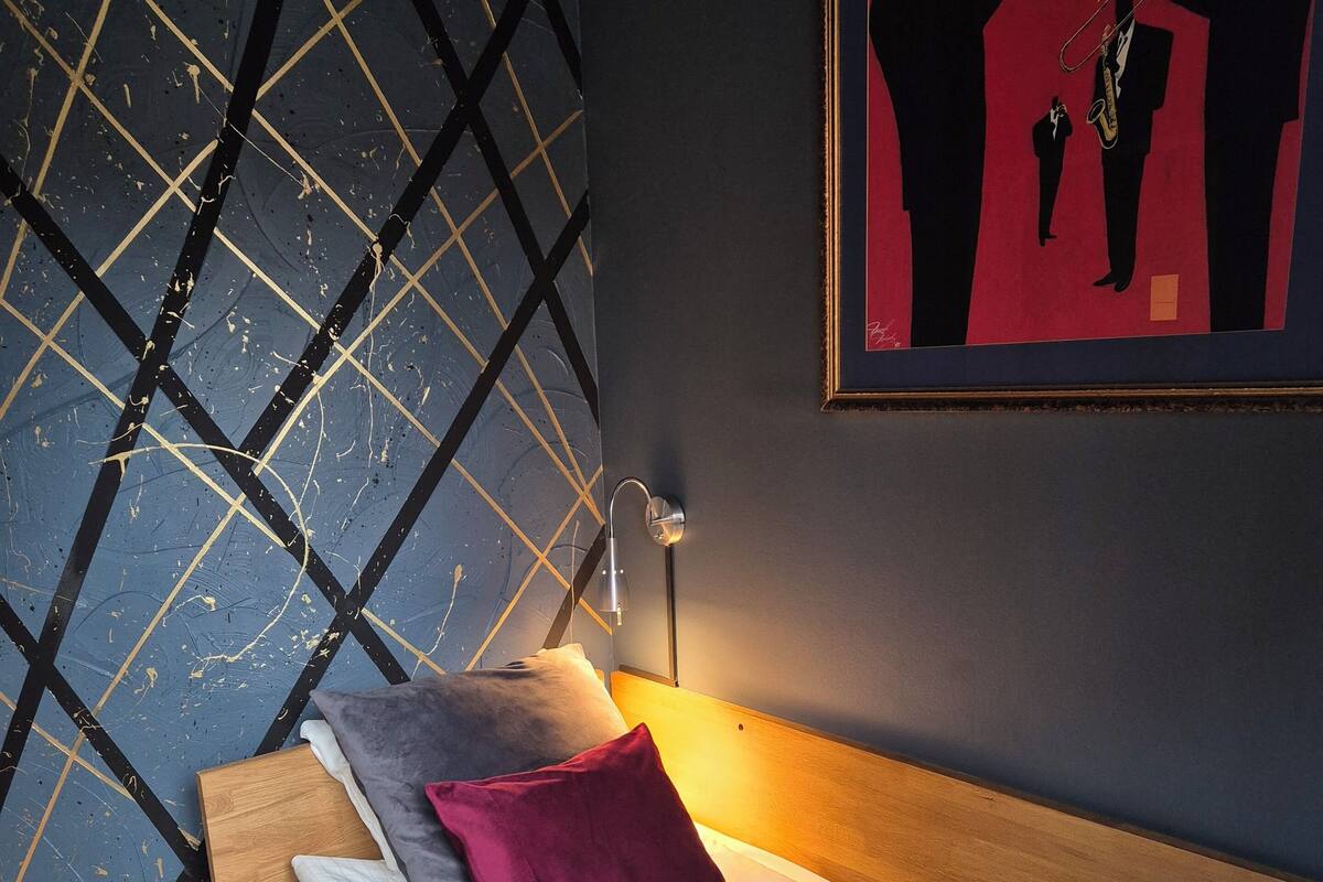 Een slaapkamer met een bed, kussens, een nachtkastje, een wandlamp en een lijst met een afbeelding.