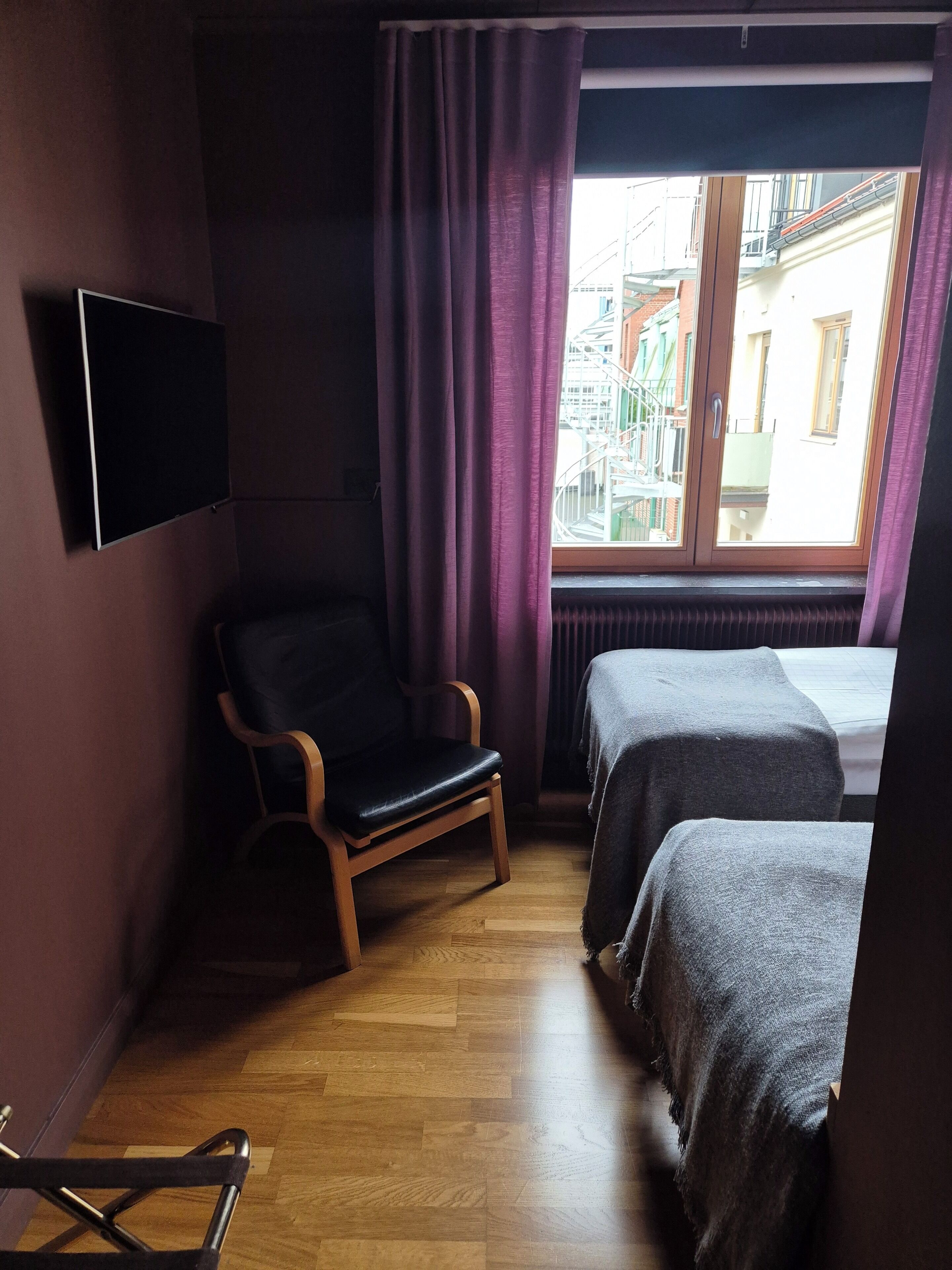 Ett litet hotellrum med trägolv, en enkel stol, två sängar, en tv som är monterad på väggen och ett fönster med gardiner.