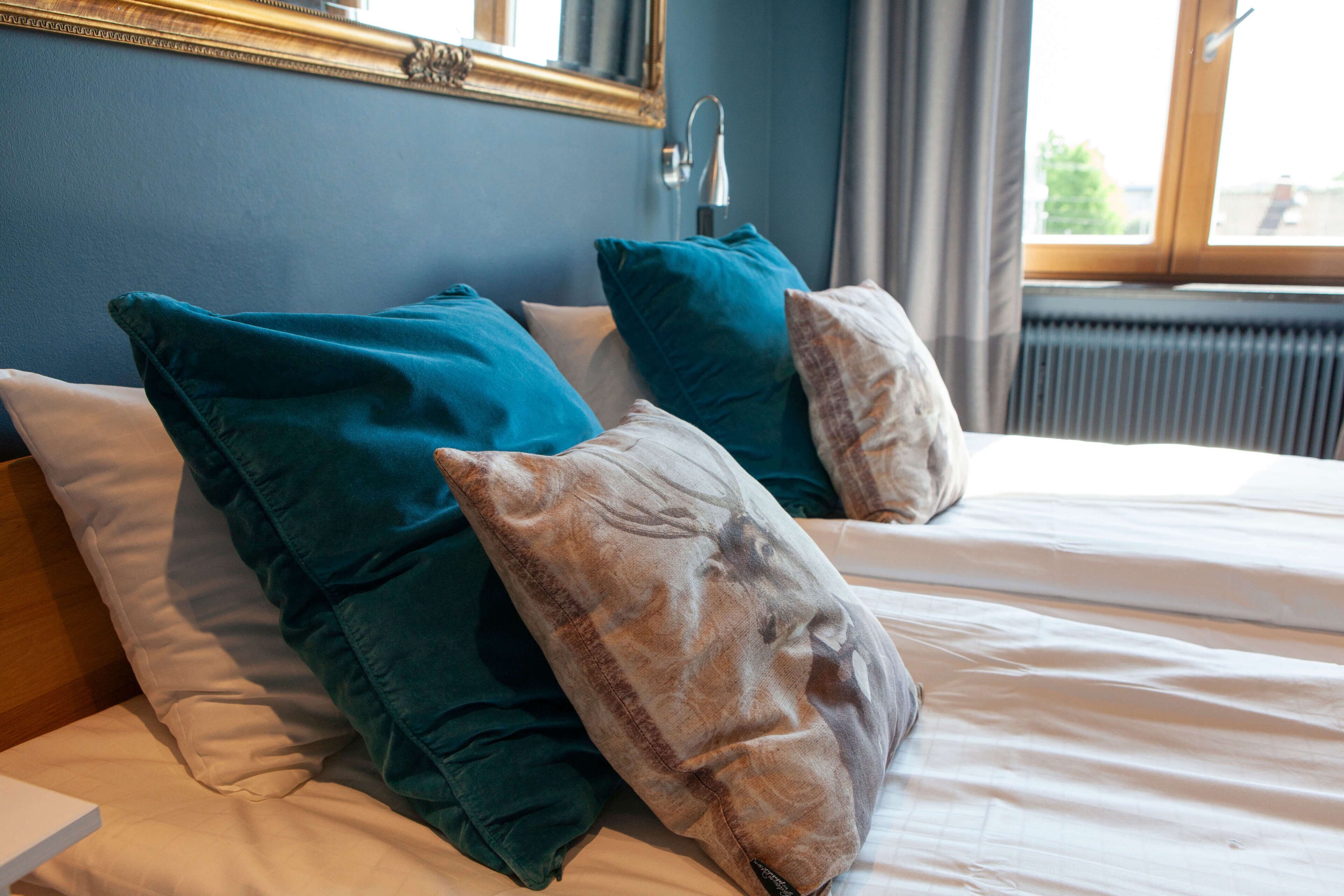 A bed with teal and beige pillows, a wooden headboard, a mirror above the bed, and a window with curtains.