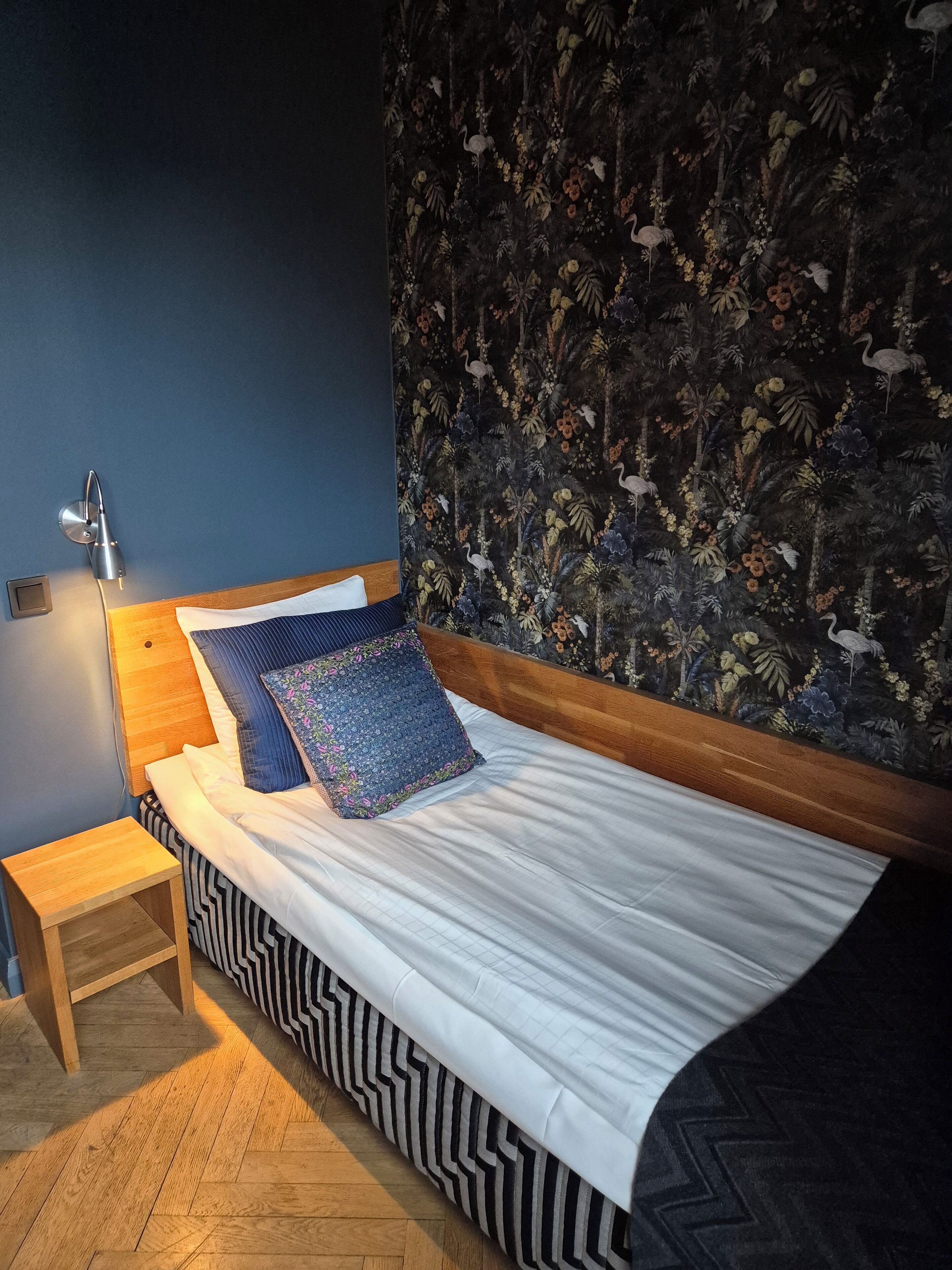 A bedroom with a bed, a nightstand, a wall-mounted lamp, and a floral wallpaper.