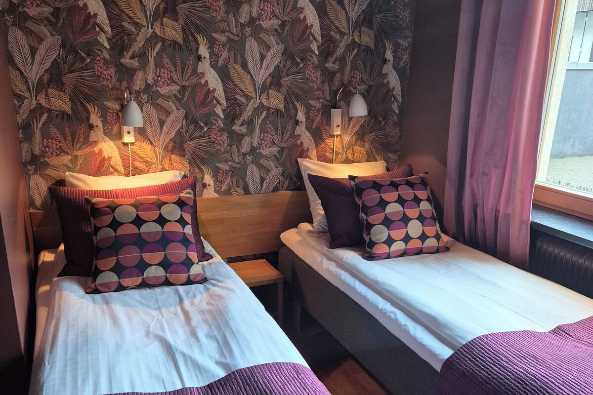 Een kamer met twee bedden, elk voorzien van een gekleurd kussen en een lamp. De muren zijn bekleed met behang met een bladerpatroon.