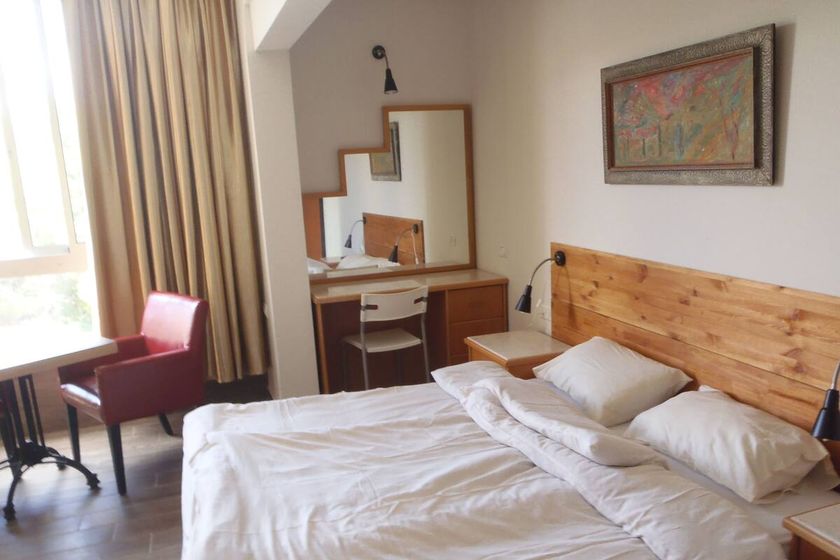 A hotel room with a bed, a desk, a chair, a mirror, and a painting on the wall.