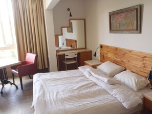 A hotel room with a bed, a desk, a chair, a mirror, and a painting on the wall. - Marom Carmel Center Apartments (Haifa)