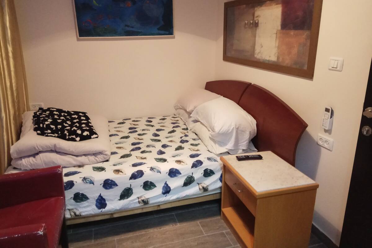 A small, neatly arranged bedroom with a bed, a bedside table, a red chair, and two paintings on the walls.