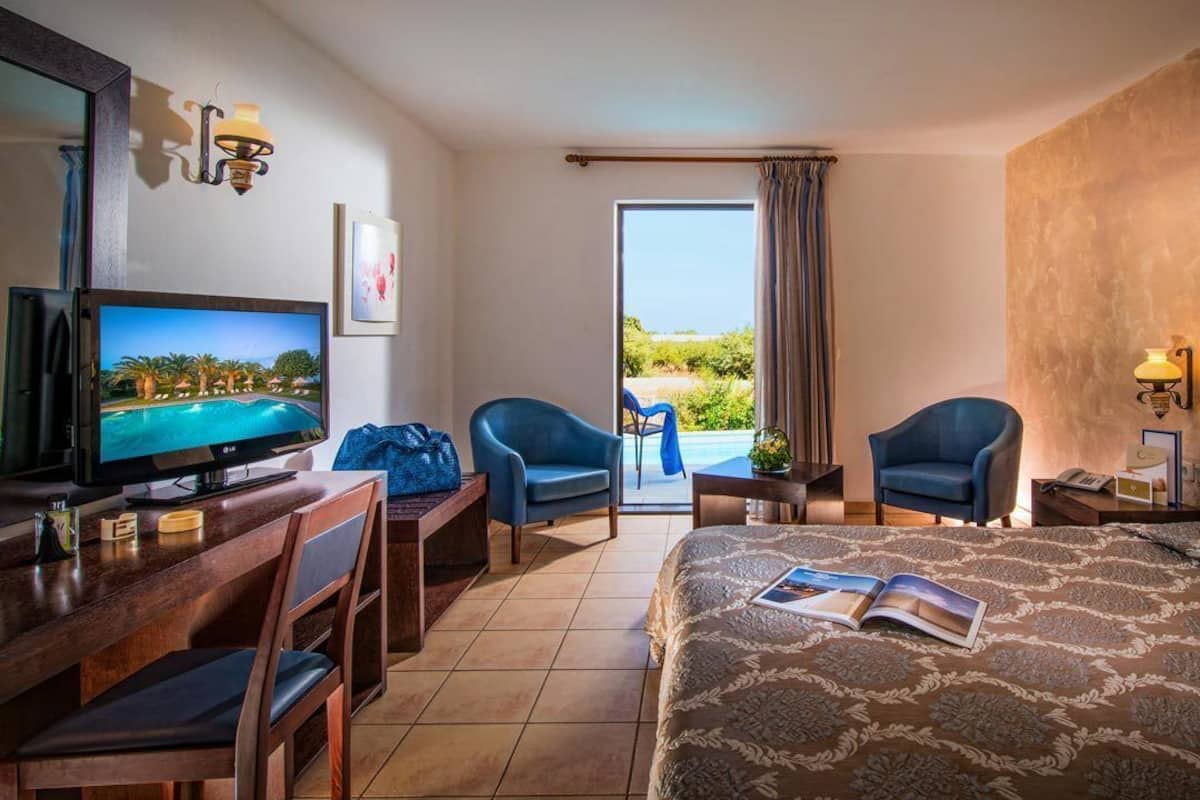 A hotel room with a bed, a TV, two chairs, a small table, and a view of a pool.