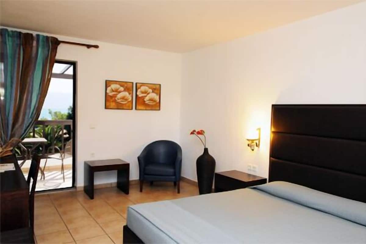 A hotel room with a bed, a chair, a small table, a vase with flowers, and two wall-mounted lamps.