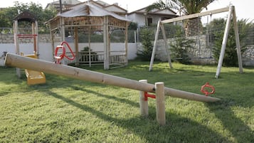 Une balançoire à bascule avec une structure en bois et des poignées rouges, installée sur une pelouse verdoyante, avec une maison et un espace de jeux clôturé en arrière-plan.