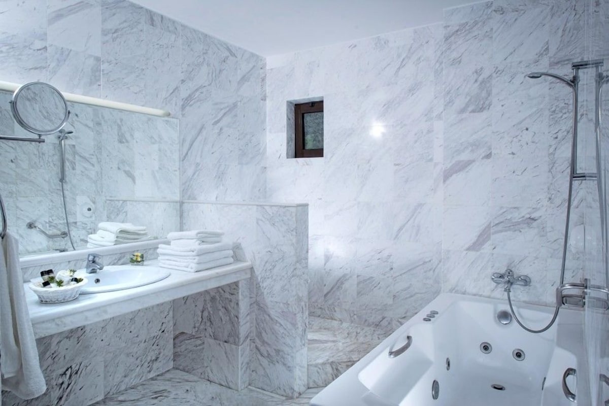 A bathroom with a marble wall, a sink with a mirror, a bathtub, and a small window.