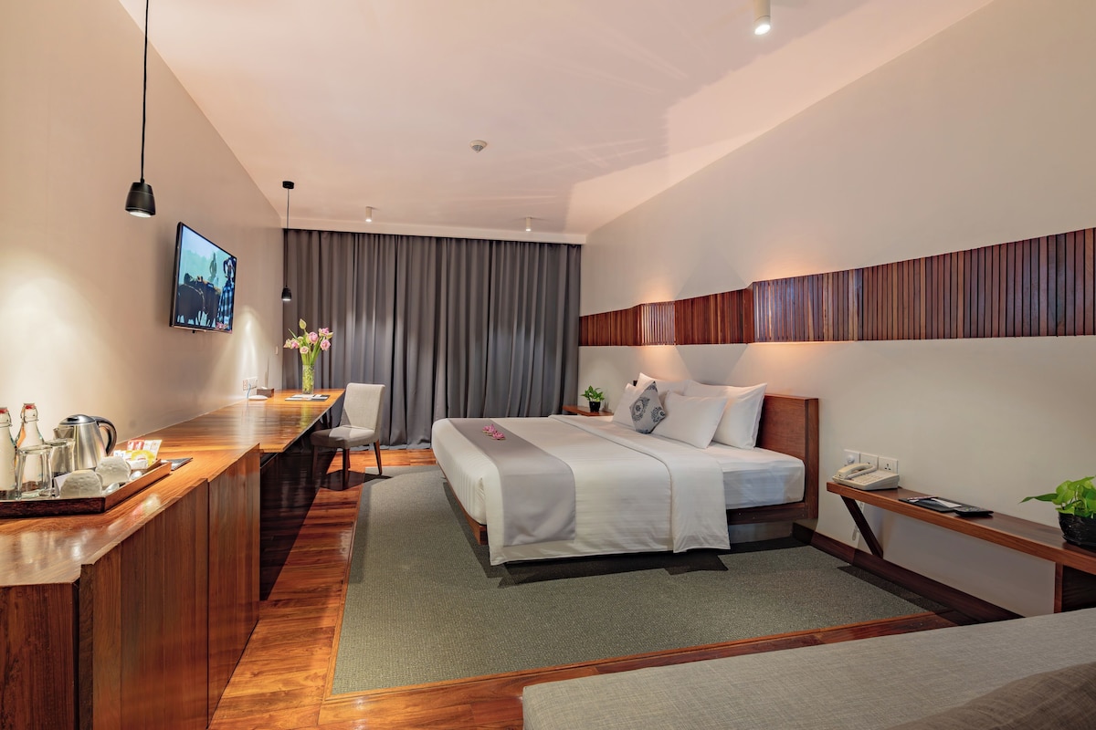 Une chambre d’hôtel moderne avec un bureau en bois, un lit recouvert de draps blancs, une table de chevet avec un téléphone et un téléviseur fixé au mur.