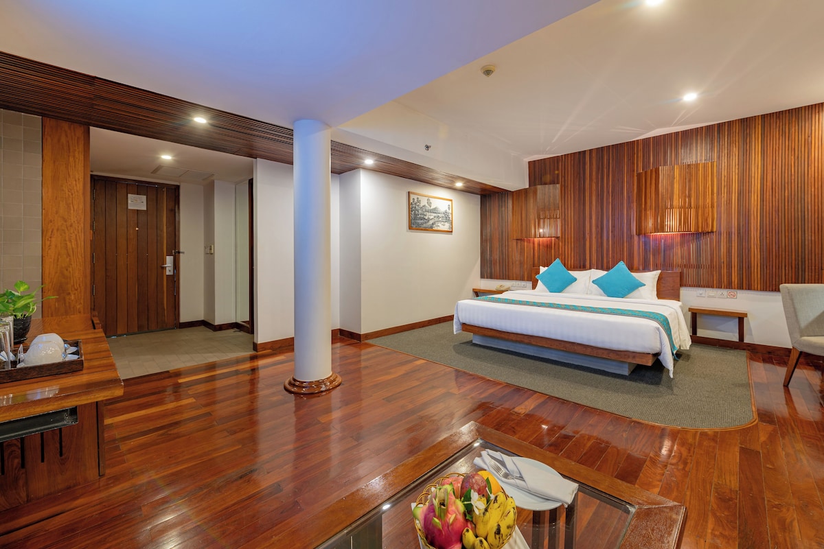 Une chambre d’hôtel avec un plancher en bois, un lit avec une literie blanche et des oreillers bleus, un bureau avec une chaise, et une petite table avec des fruits et un vase.