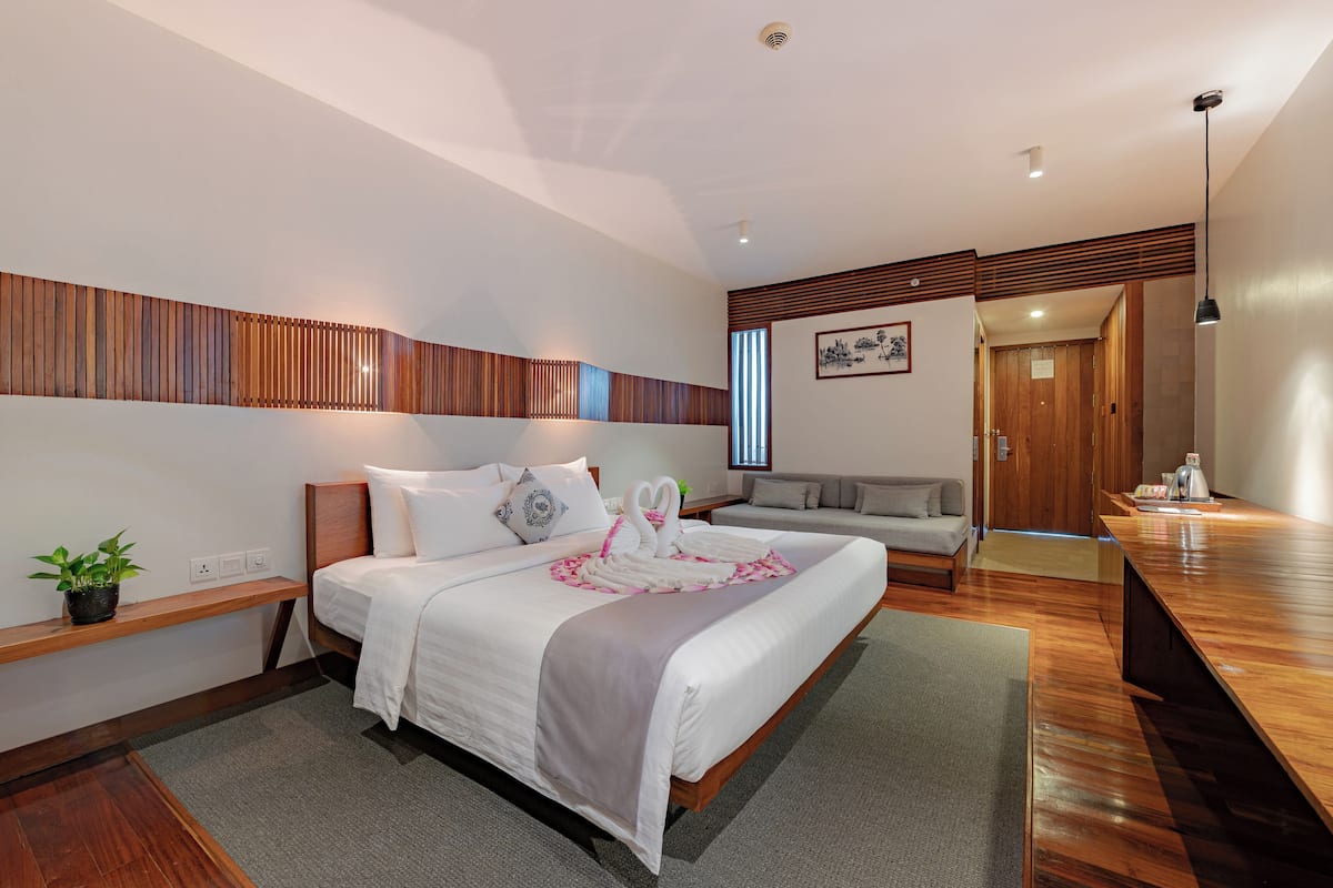 Een moderne hotelkamer met een groot bed, een houten hoofdeinde, een nachtkastje met een plant en een zithoek met een bank en een kleine tafel.