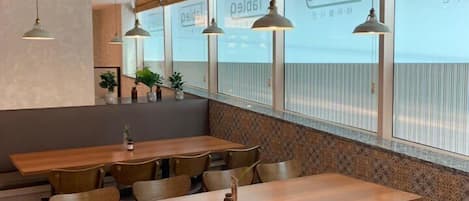 A modern dining area with wooden tables and chairs, pendant lighting, and large windows with signage.