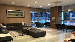 Un área de estar moderna con un sofá modular, una mesita de centro y una mesa de comedor con sillas. Amplios ventanales ofrecen vistas de la ciudad.
