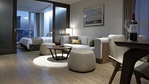 Una habitación de hotel moderna con sofá, reposapiés, mesa de centro y mesa de comedor. Se ve una cama con vista a la ciudad a través de amplias ventanas.