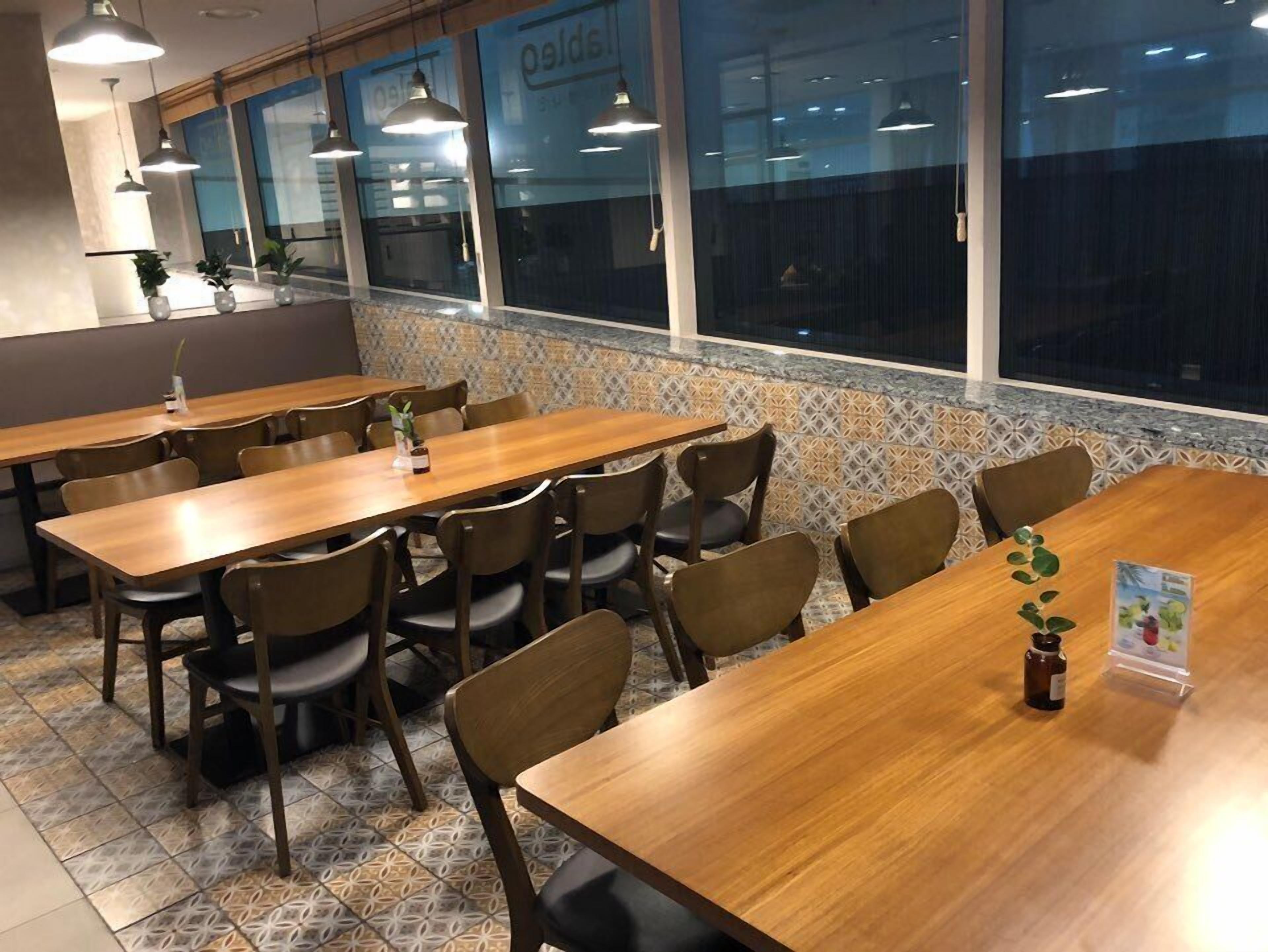 A restaurant with multiple wooden tables and chairs, large windows, and hanging pendant lights.