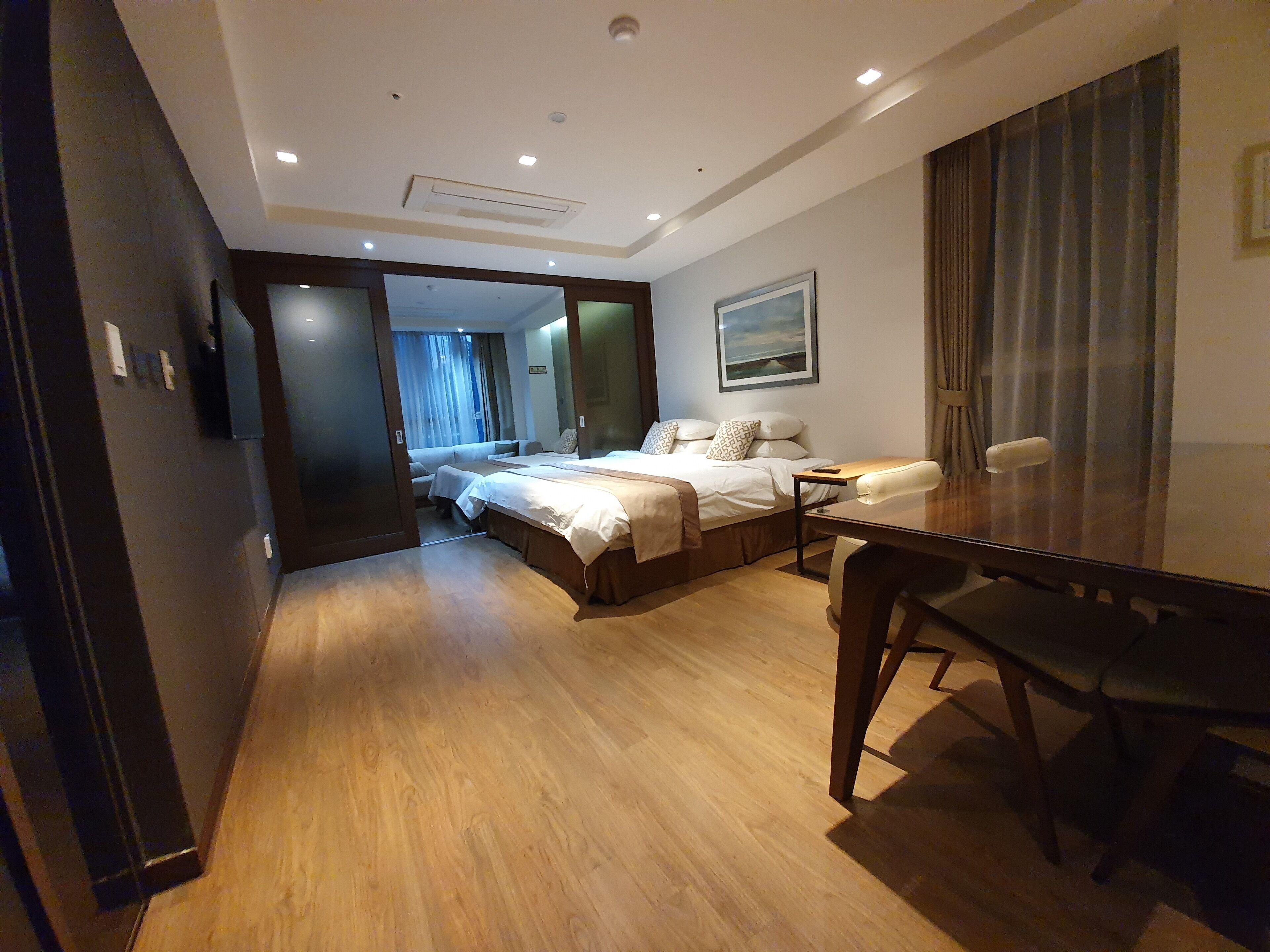A hotel room with a large bed, a desk, and a chair. There is a painting on the wall and a window with curtains.