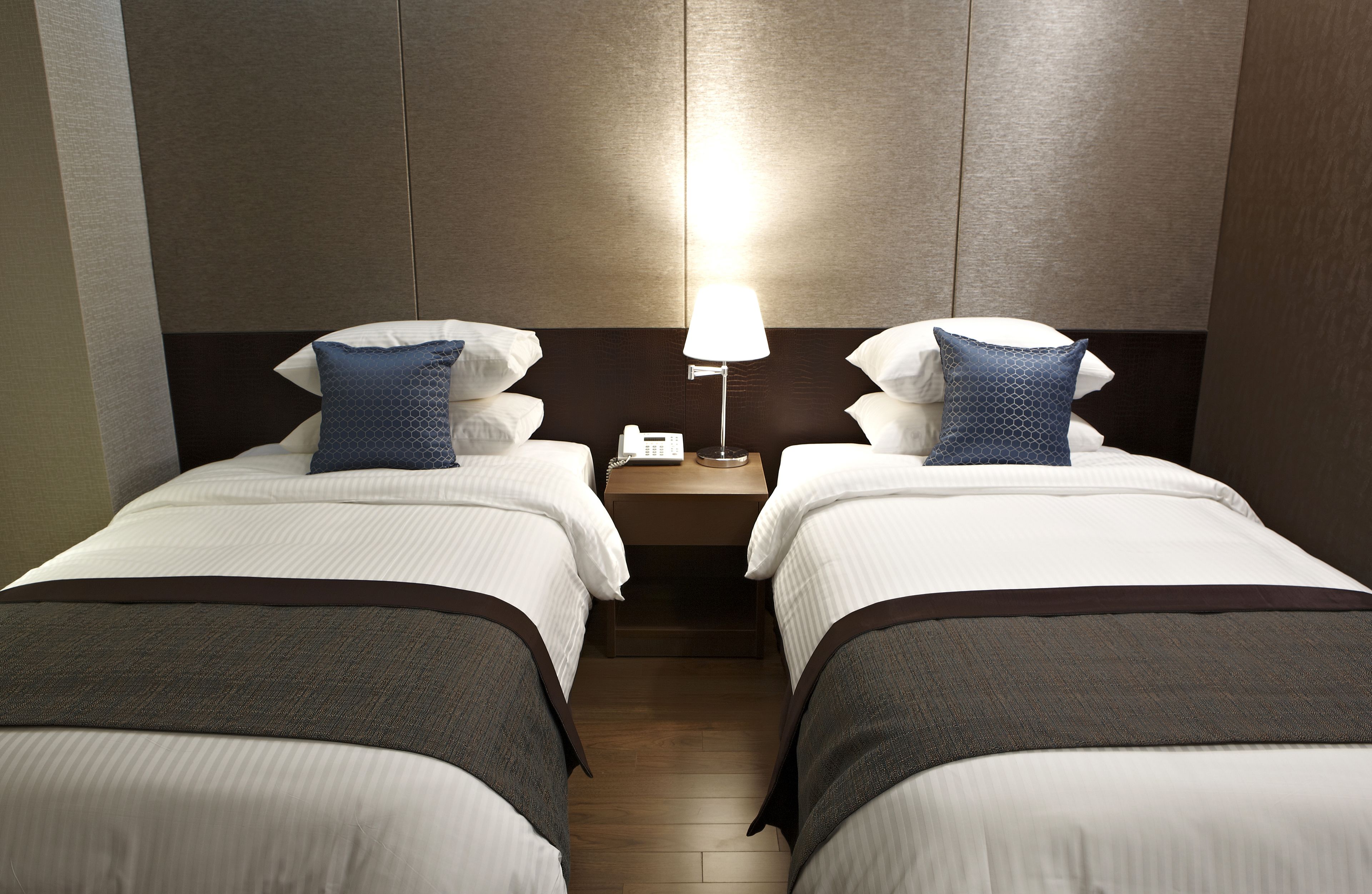 A hotel room with two single beds, each with a pillow and a bedside table with a lamp.