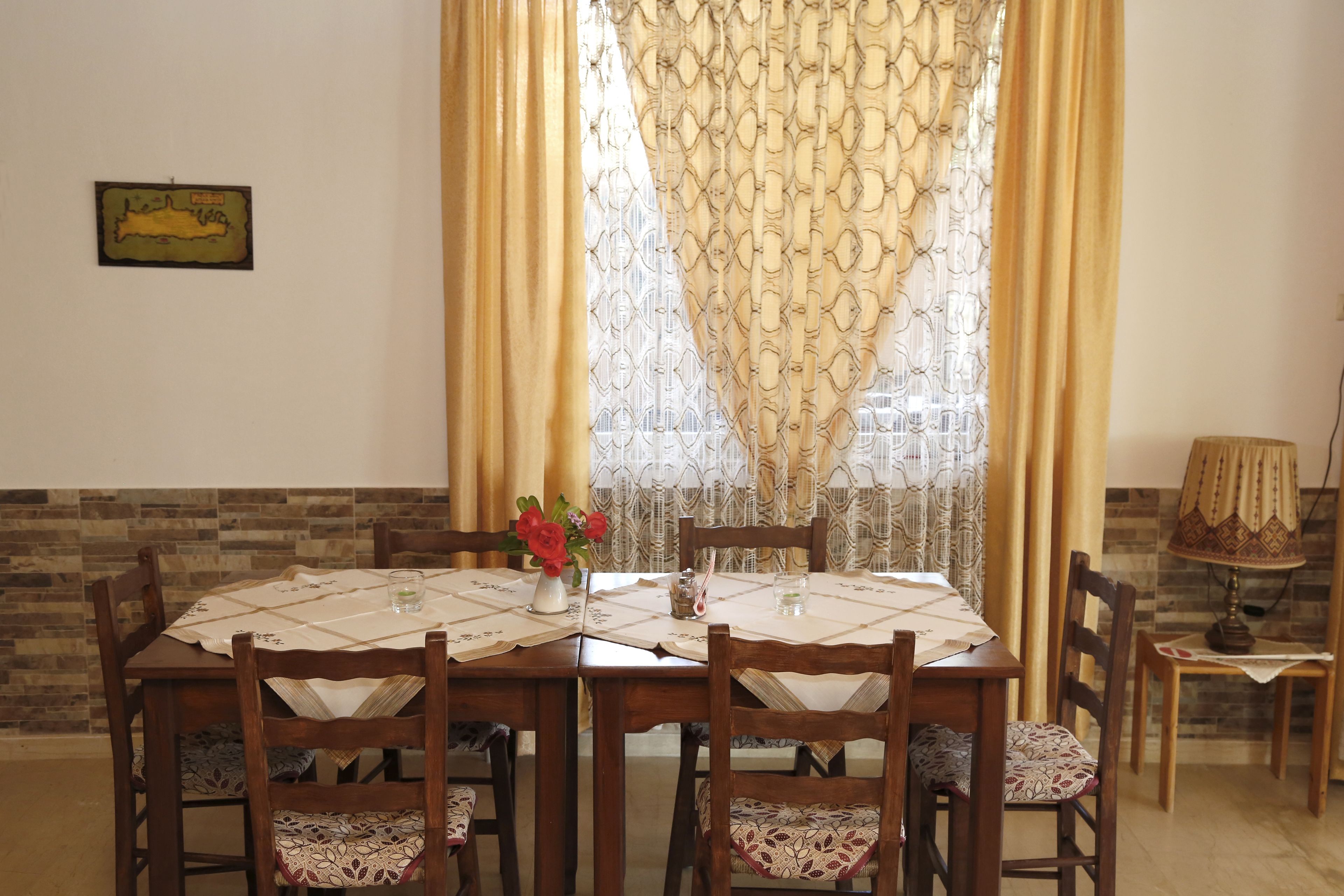 A dining area with a table set for four, surrounded by wooden chairs with patterned cushions. A vase with flowers is on the table, and a lamp is on a side table.