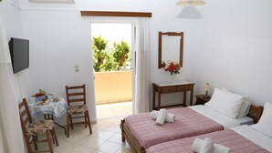 A hotel room with two beds, a dining table with chairs, a TV, and a balcony with a view of greenery. - Athina Rooms (Rethymno)