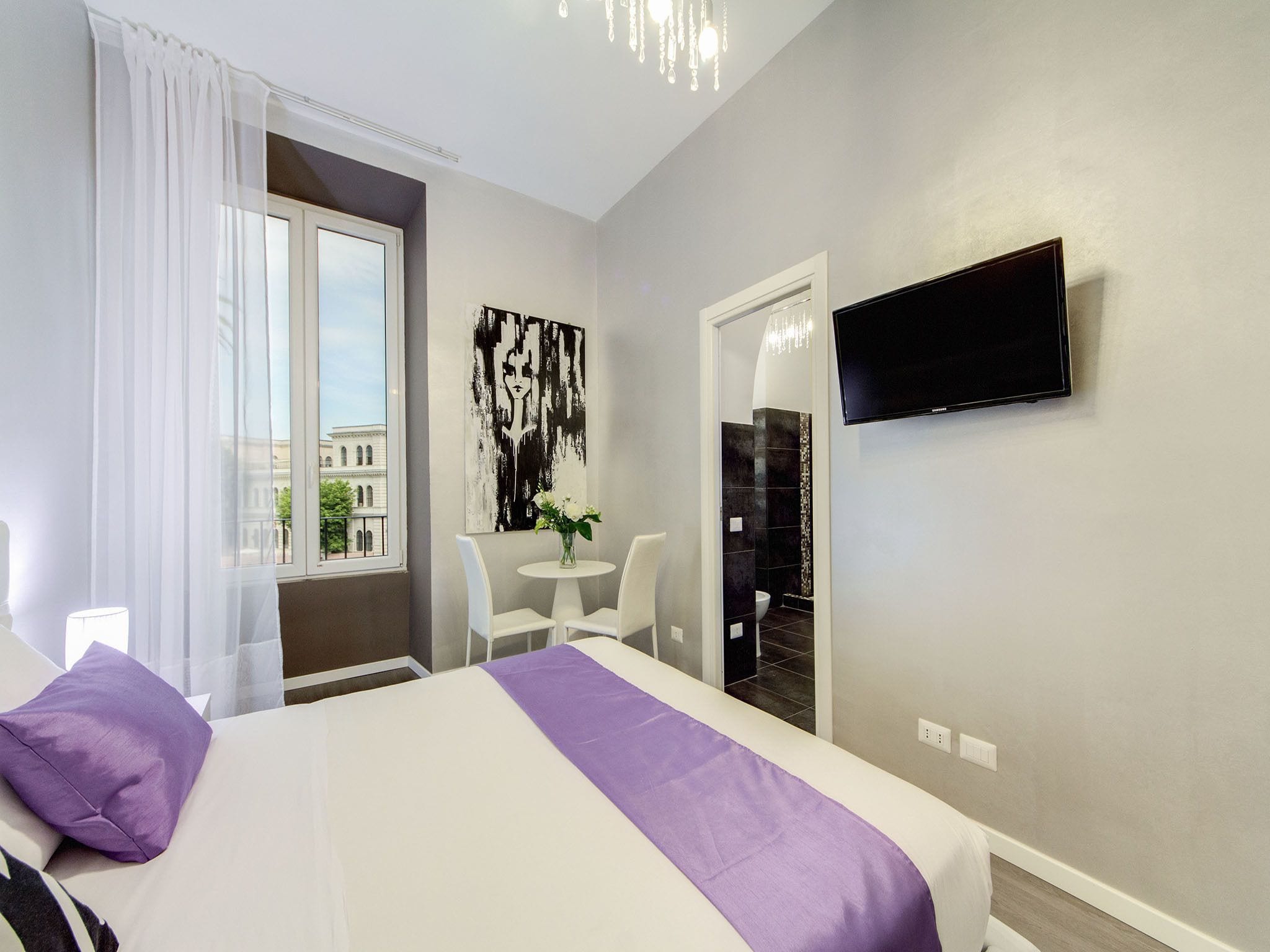 A hotel room with a bed, a TV, a small table with chairs, and a window with curtains.