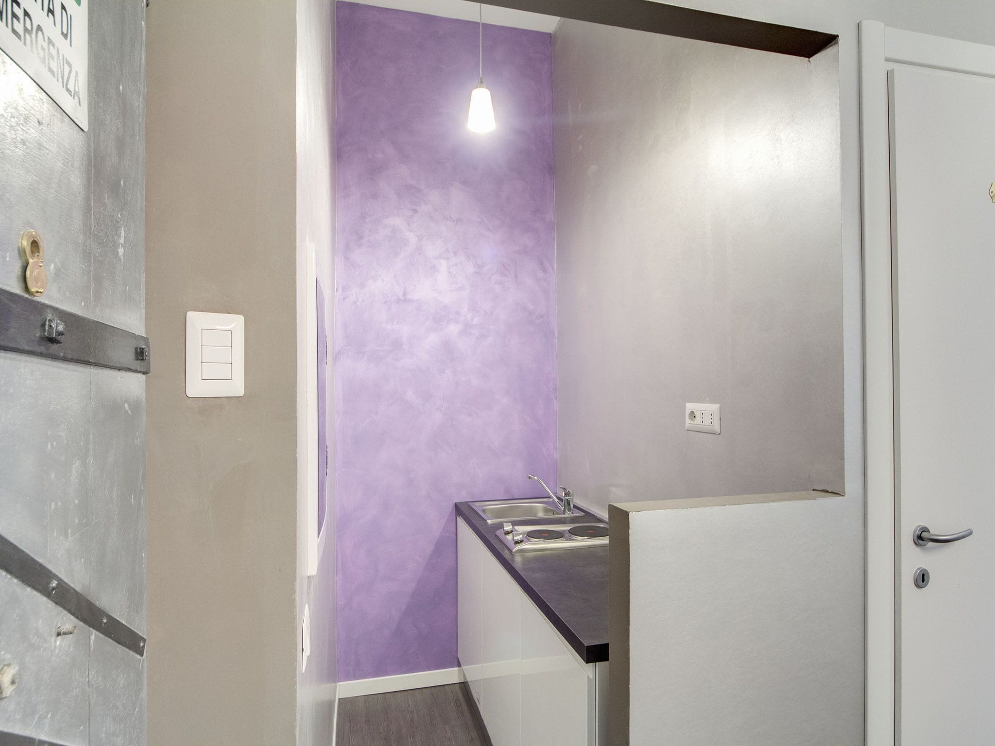 Une petite cuisine équipée avec un mur d’accent violet, un luminaire et une porte dotée d’une poignée.