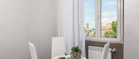 Un pequeño balcón con una mesa y sillas blancas, una planta en maceta y vistas a un paisaje urbano a través de una ventana.