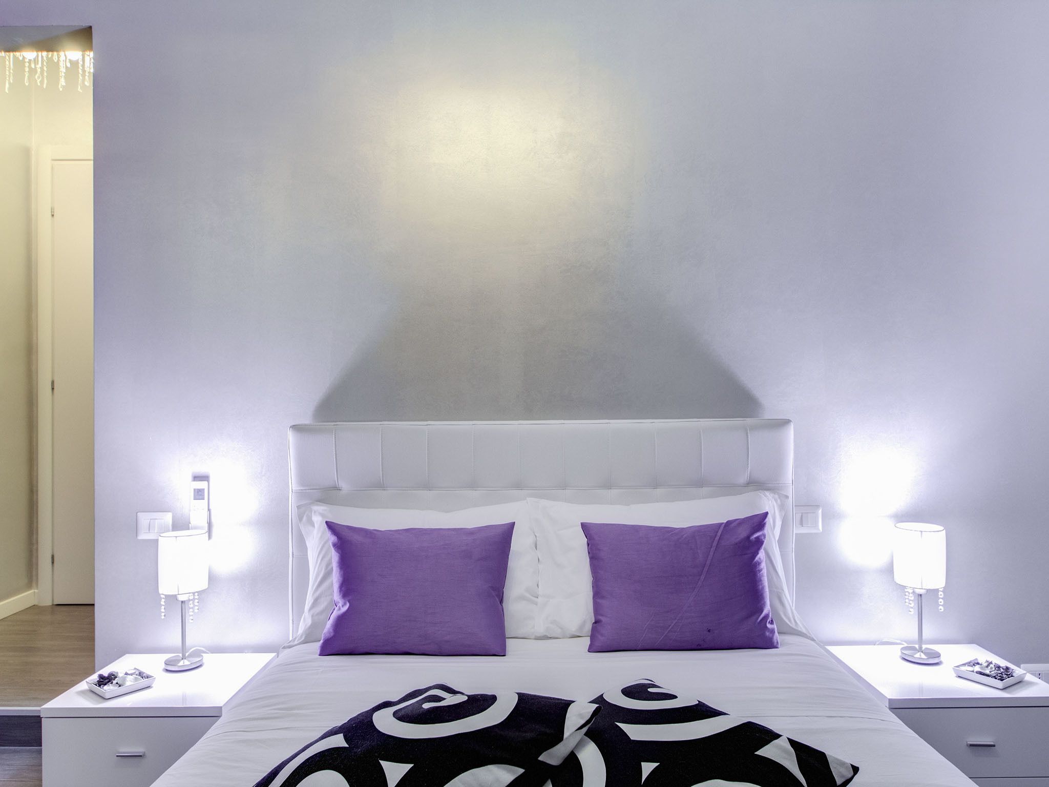 A neatly made bed with a white headboard and two purple pillows, flanked by two bedside tables with lamps and a small tray of fruits.