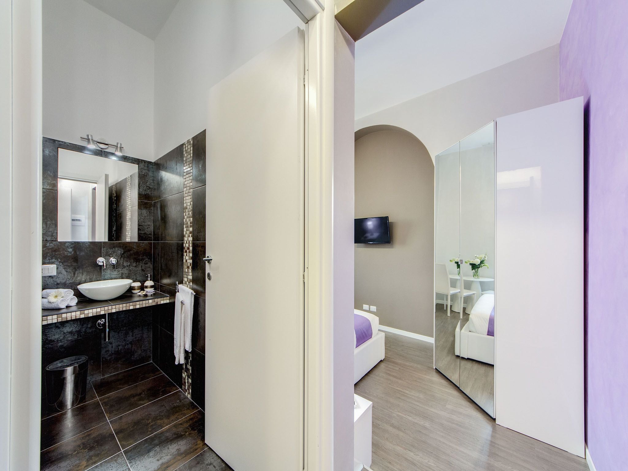 Un bany modern amb un mirall gran, un lavabo i un vàter. Hi ha un televisor penjat a la paret i una taula petita amb un ram de flors.