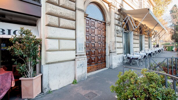 Un edificio con una puerta de madera grande, asientos exteriores con mesas y sillas, y una planta en maceta cerca de la entrada.