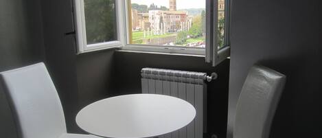 A room with a round table and two white chairs, a window with a view of buildings, and a radiator.