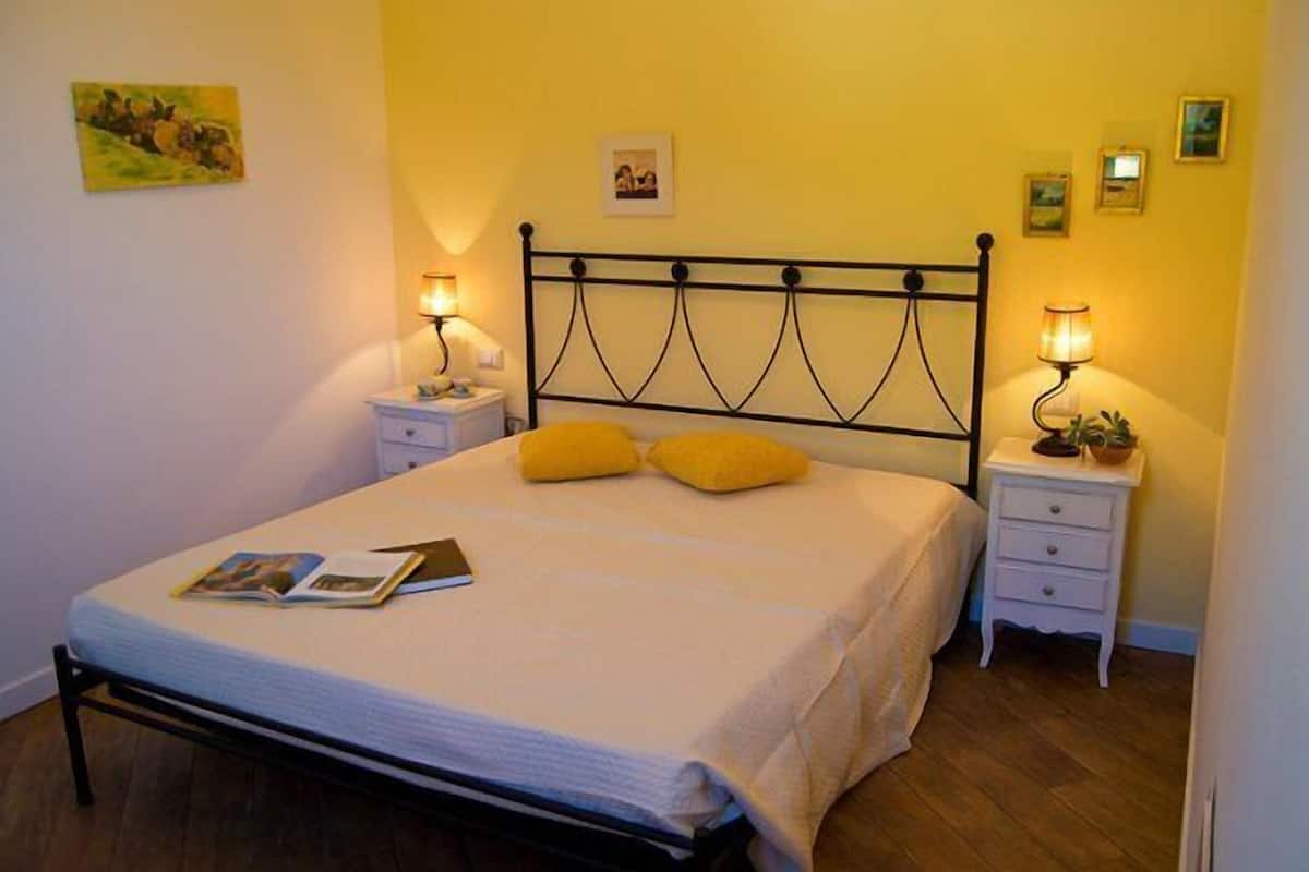 Un lit bien fait, avec un matelas blanc et deux oreillers, flanqué de deux tables de chevet surmontées de lampes, dans une pièce aux murs jaunes et au plancher en bois.