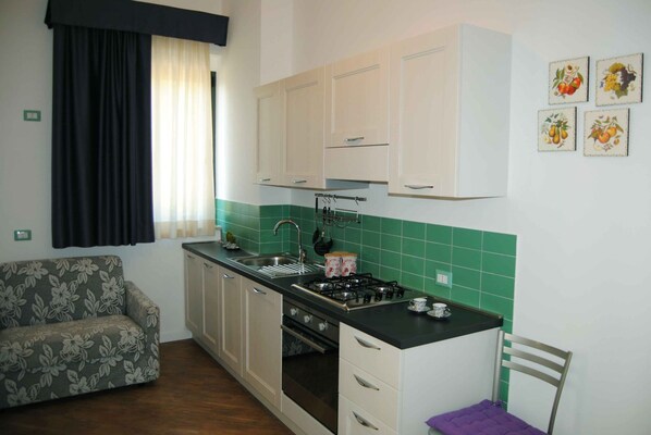 Cocina compacta con estufa a gas, lavamanos y armarios. En la zona de estar contigua hay un sofá y una silla.