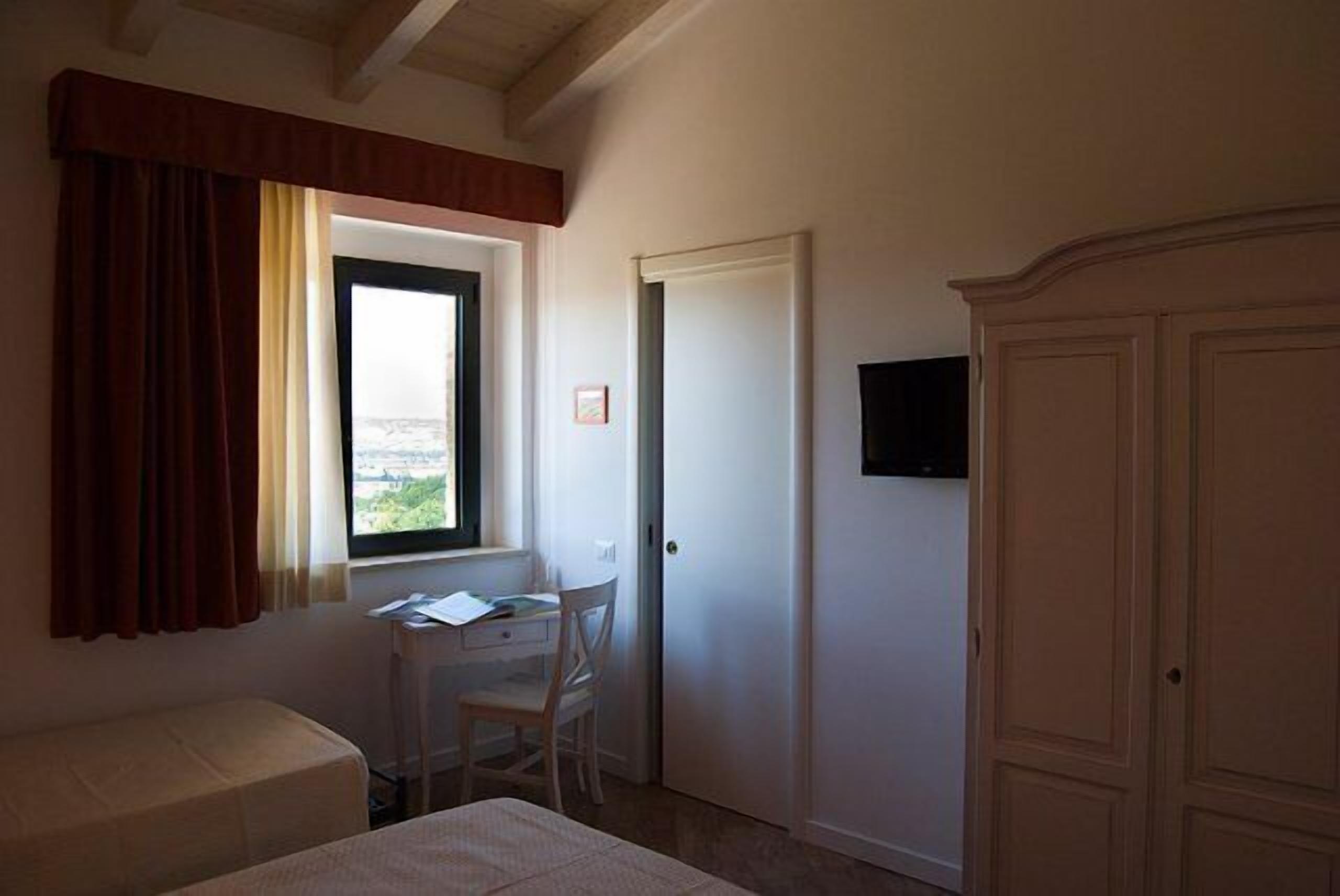 A bedroom with a bed, a desk, and a chair. There is a window with curtains, a door, and a wardrobe.