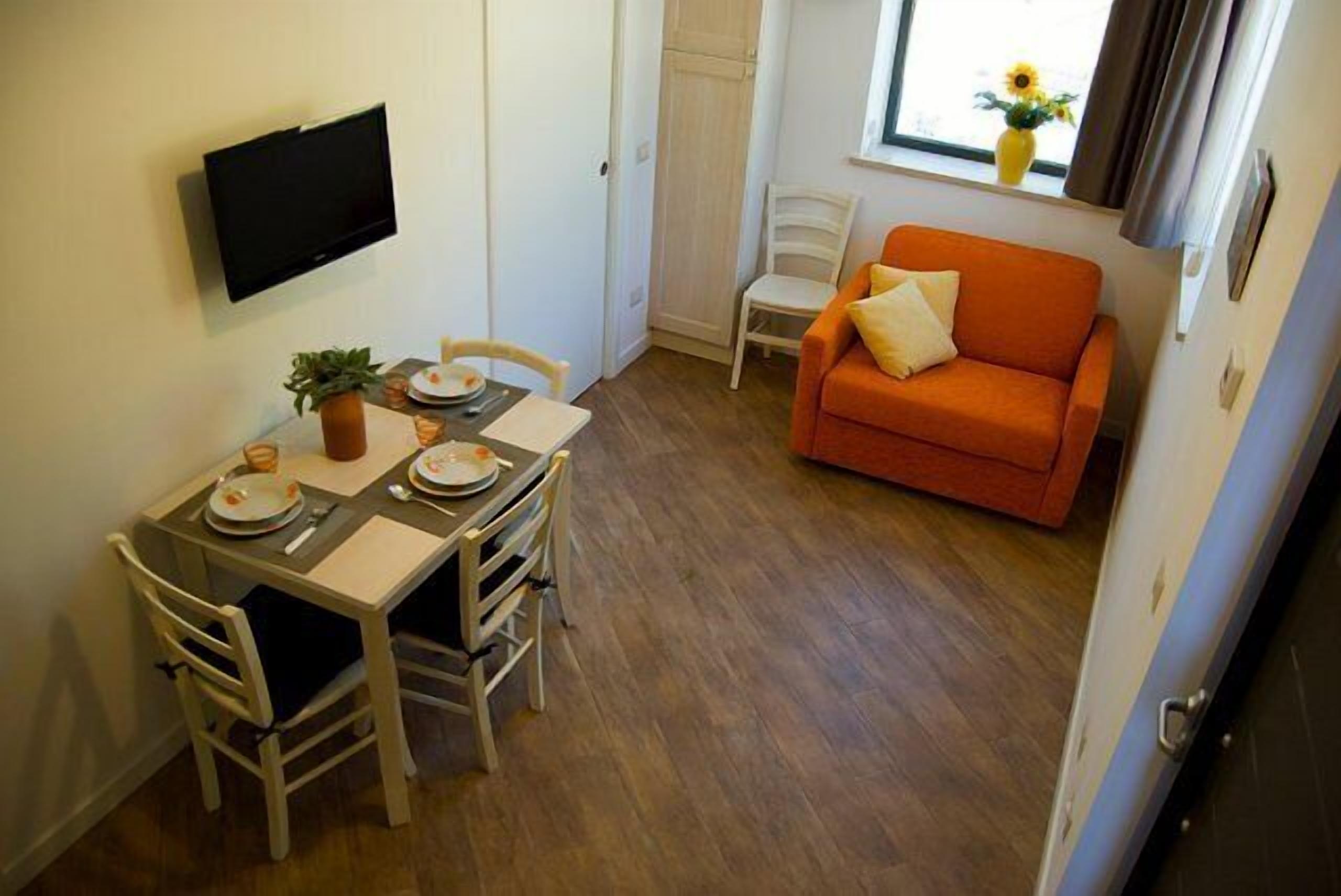 A compact living space with a dining table set for two, a sofa, a TV mounted on the wall, and a window with a view of the outdoors.