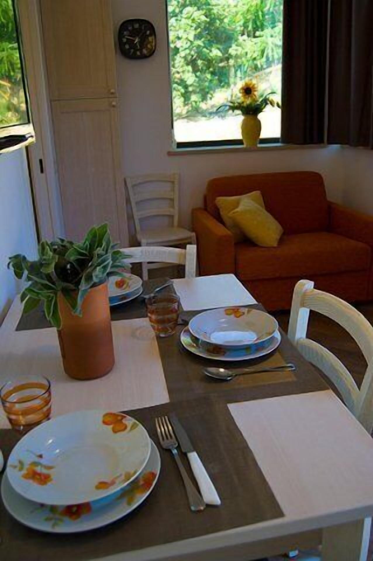 Una tavola imbandita con piatti, posate e un vaso di fiori. Si vede una finestra con vista su del verde e un orologio appeso al muro.