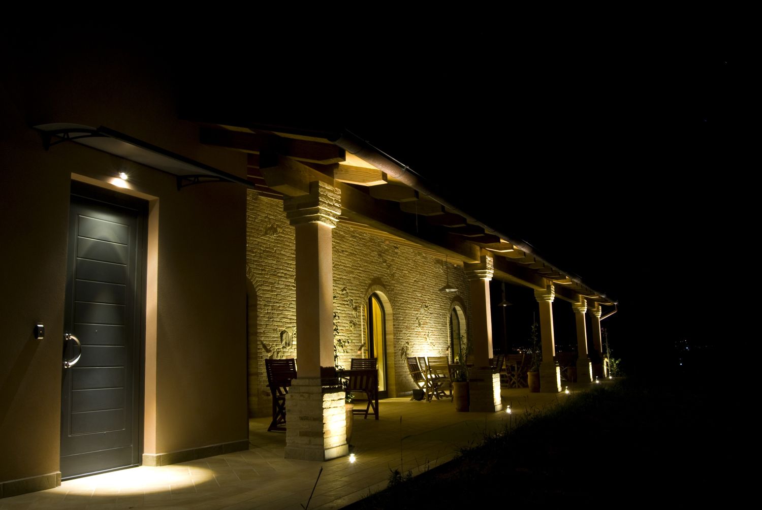 A building with a stone facade, a dark entrance door, and a covered outdoor seating area with chairs and tables.