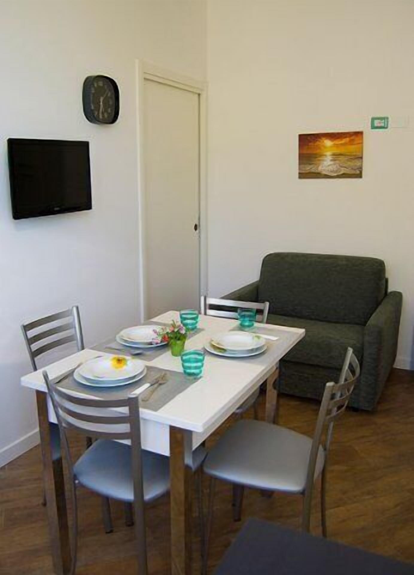 Un comedor con una mesa blanca dispuesta para cuatro personas, dos sillas a cada lado, un sofá verde, un reloj y un cuadro en la pared.
