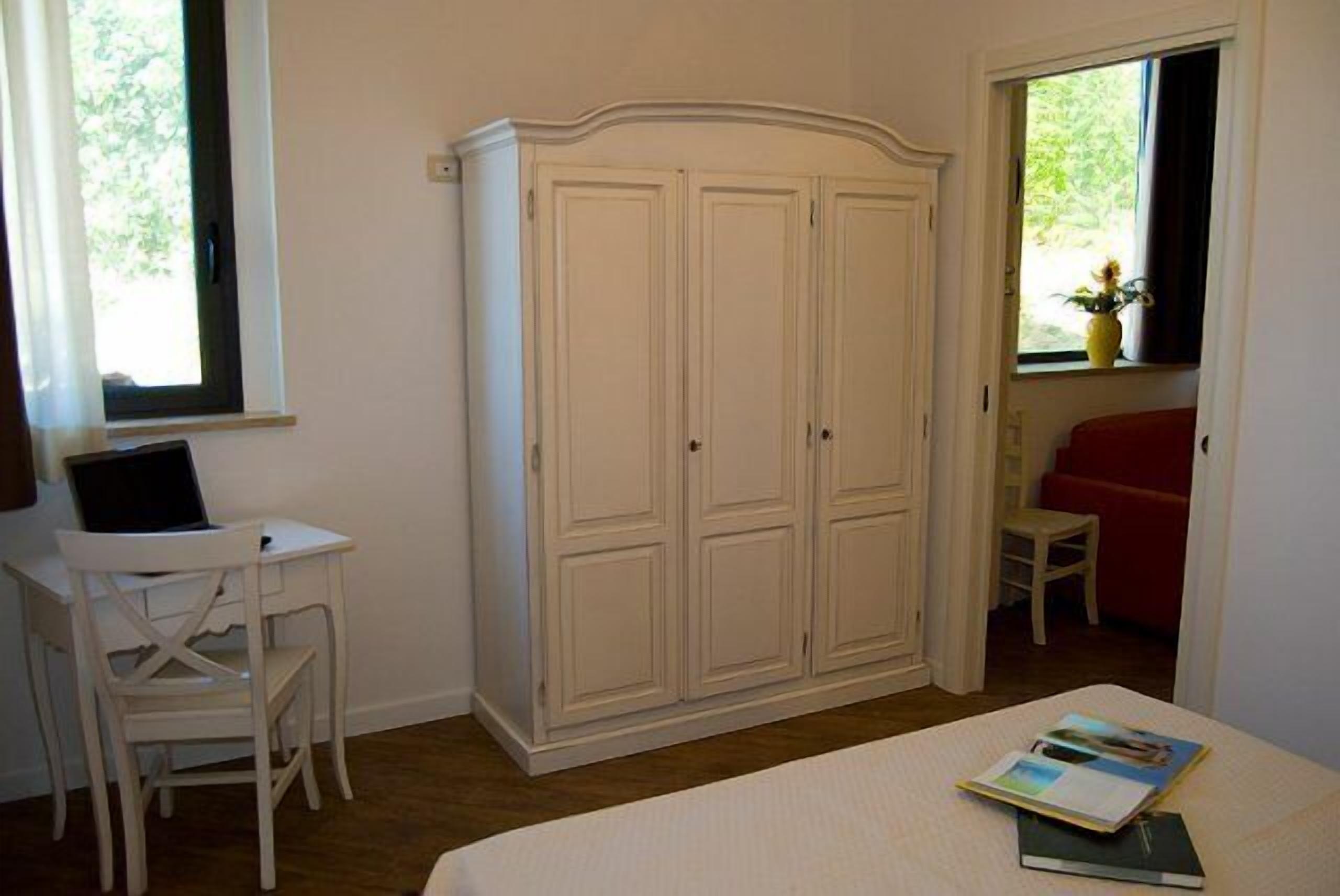 Ein Schlafzimmer mit Bett, Schreibtisch mit Laptop, Kleiderschrank und Fenster mit Blick auf Grünflächen.