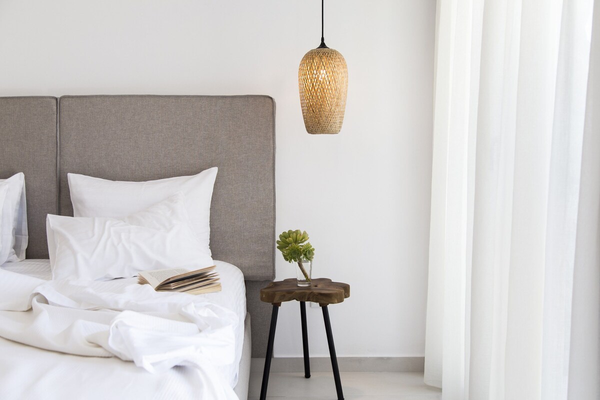 A neatly made bed with white linens, a small bedside table with a plant, and a hanging woven light fixture.