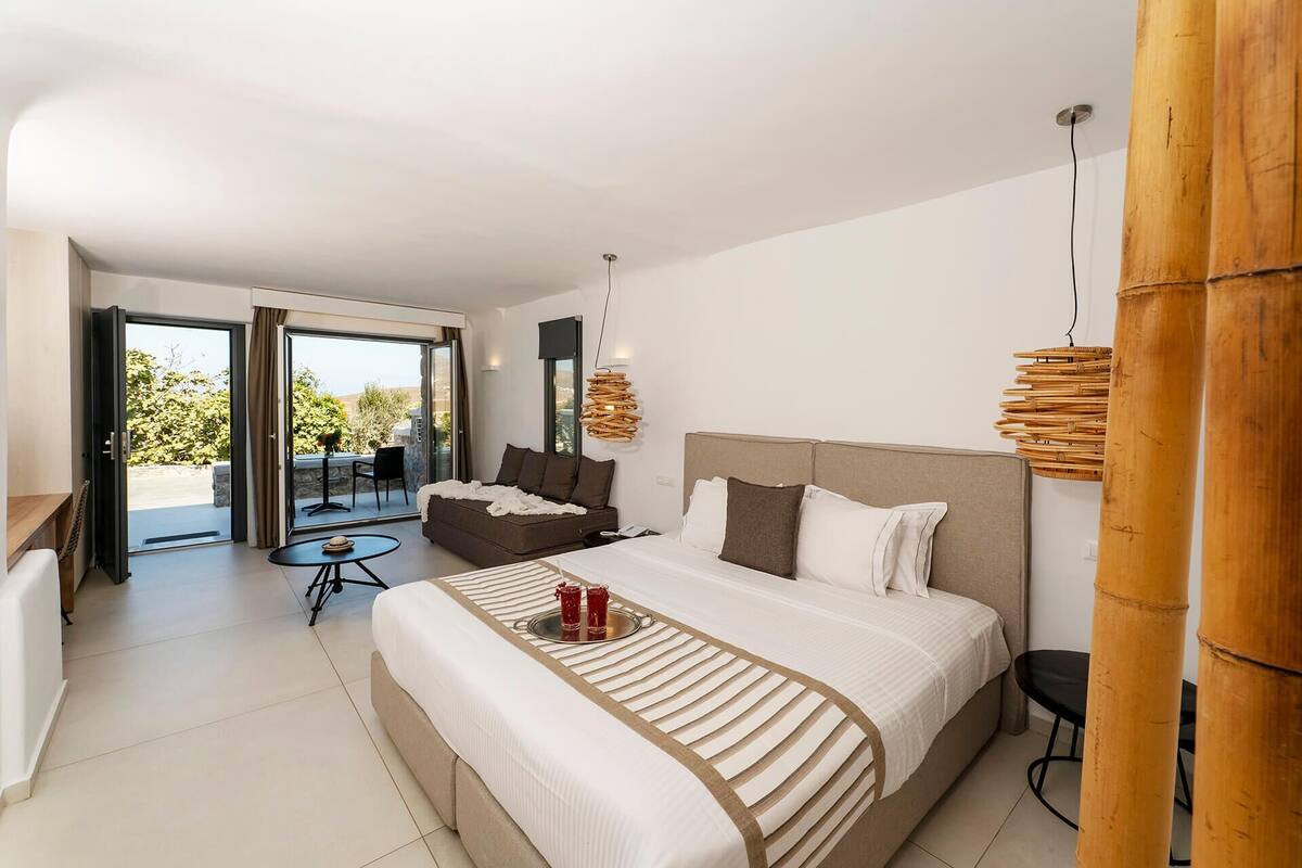 A modern hotel room with a large bed, a balcony with a table and chairs, and a view of the outdoors.