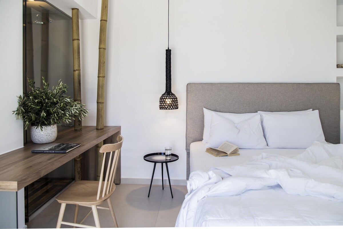 Une chambre d’hôtel moderne, équipée d’un lit, d’une table de chevet, d’une chaise, d’une petite table avec un verre et d’un luminaire suspendu.