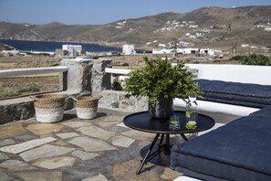 Una terraza con suelo de piedra, una mesa redonda con planta en maceta, cestas de mimbre y un vaso de bebida.
