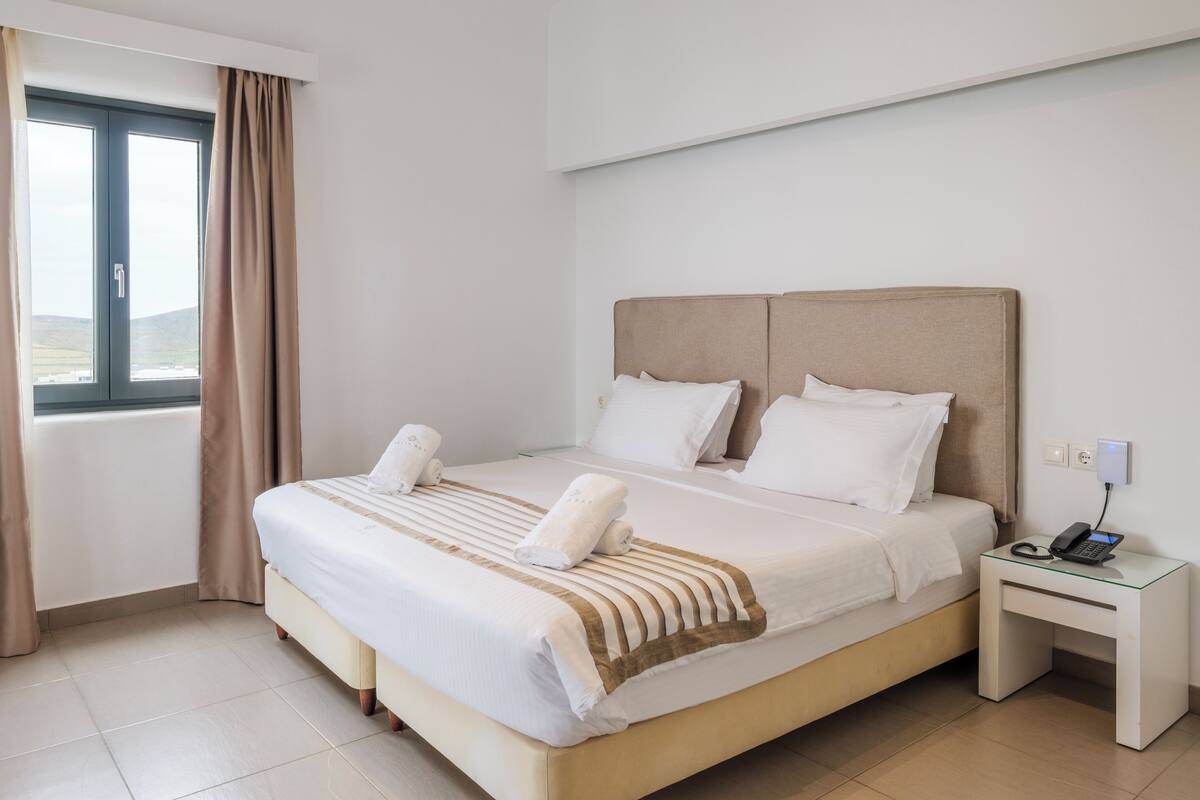 A hotel room with a bed, a bedside table with a phone, and a window with curtains.