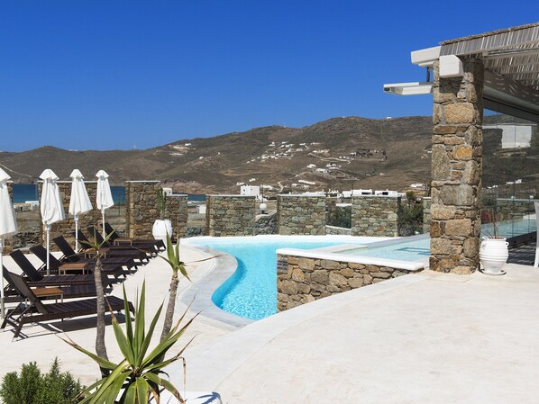 Zona de piscina con sillones de descanso y sombrillas, paredes de piedra y vistas a las montañas y al mar.